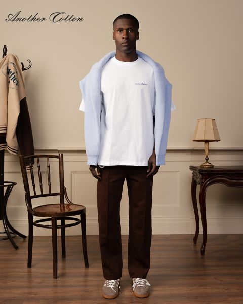 Una persona è in una stanza, indossa un maglione azzurro sulle spalle, una maglietta bianca e pantaloni marroni. Nella stanza ci sono una sedia di legno, una lampada da terra vintage e una giacca su un appendiabiti. Nell'immagine si vede il testo "Another Cotton".