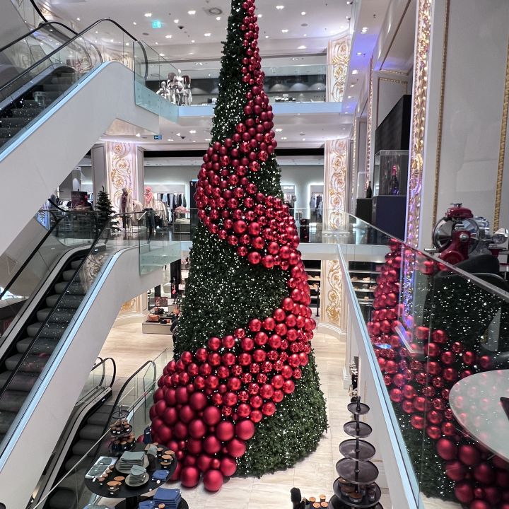 Un grande albero di Natale in un centro commerciale a più piani è decorato con palline rosse e una spirale di decorazioni floreali rosse. Intorno all'albero si vedono acquirenti e scale mobili, il che contribuisce all'atmosfera festosa.