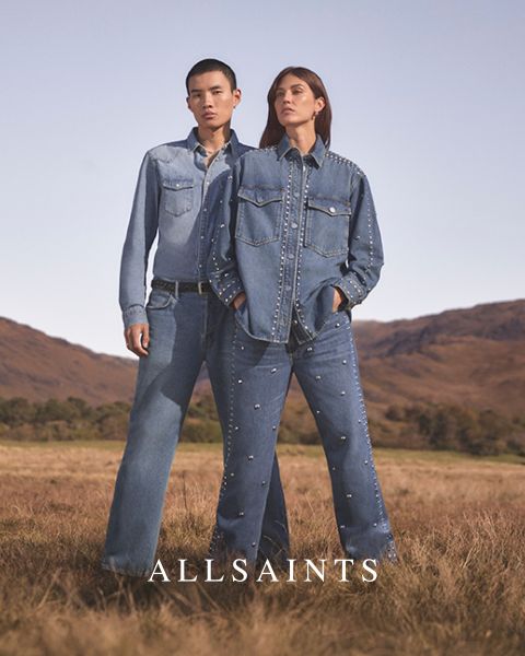 Due persone sono in piedi in un campo aperto e indossano abiti di jeans decorati con borchie. La persona a sinistra indossa una camicia di jeans chiara e jeans, mentre la persona a destra indossa una giacca di jeans scura e jeans. In basso c'è scritto "ALLSAINTS".