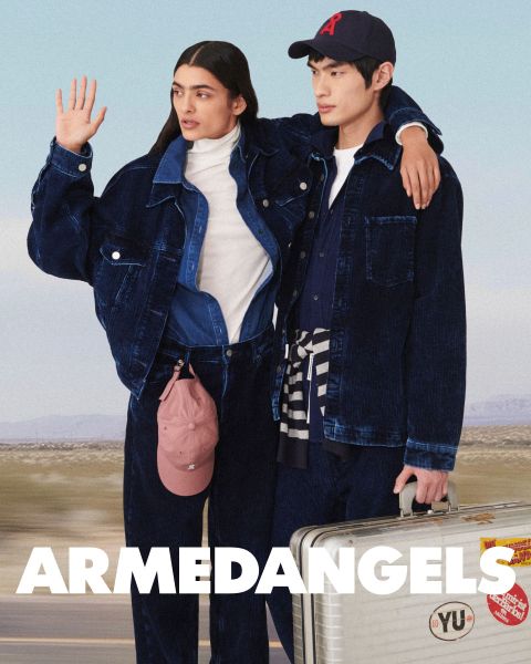 Due giovani adulti in abbigliamento di jeans stanno insieme all'aperto, uno con il braccio alzato, l'altro con una valigia coperta di adesivi. In basso, in grassetto e scritta bianca, c'è la scritta "ARMEDANGELS".