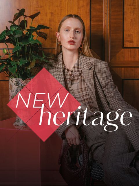 Una donna in tailleur a quadri siede accanto a una pianta, davanti a lei appare la scritta "NEW heritage".