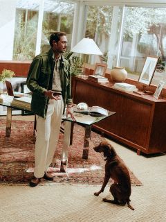 Un uomo sta in piedi con un telefono in mano accanto a una scrivania in un ufficio illuminato dal sole, mentre un cane marrone è seduto su un tappeto a motivi e lo guarda.