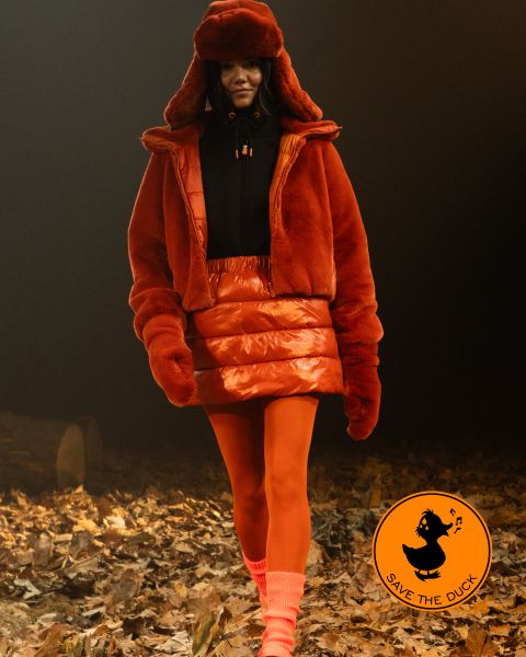 Una modella cammina su un sentiero coperto di foglie. Indossa una giacca di pelliccia arancione, una gonna abbinata e un cappello con calze arancioni e calzini color pesca. Nell'angolo si vede il logo di "Save the Duck".