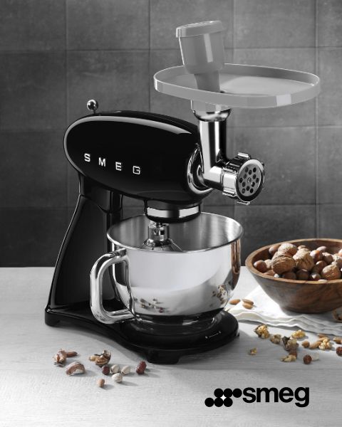 Un robot da cucina Smeg nero con accessorio tritacarne è posizionato su un bancone da cucina. Sotto c'è una ciotola in acciaio inossidabile. Noci intere e tritate sono sparse su una superficie di legno nelle vicinanze. Nell'angolo in basso a destra si vede il logo Smeg.