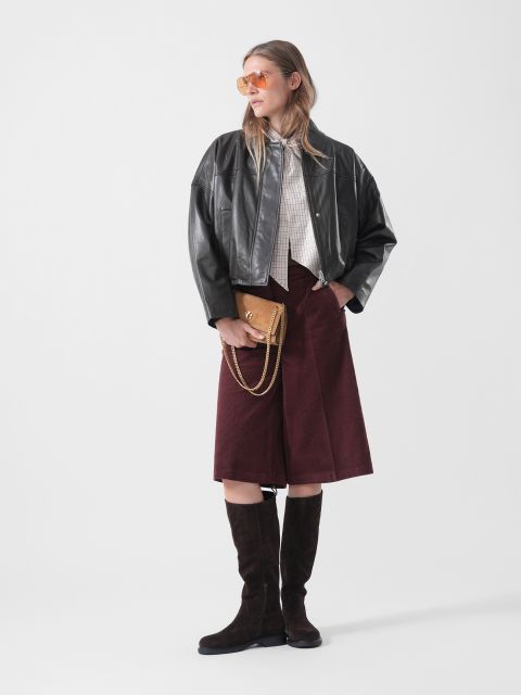 Una donna con una giacca di pelle nera, culotte bordeaux, stivali al ginocchio e occhiali da sole, che tiene una borsa marrone e sta in piedi davanti a uno sfondo bianco semplice.