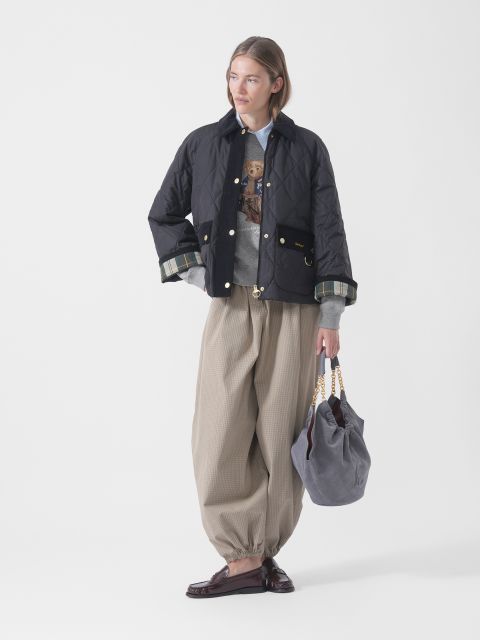 Una persona con un piumino nero, un maglione grigio, ampi pantaloni beige e mocassini tiene una borsa grigia con manici a catena dorati e si trova davanti a uno sfondo bianco.