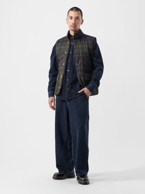 Su uno sfondo bianco semplice, una persona indossa un gilet trapuntato scuro sopra una camicia abbottonata blu navy e pantaloni larghi blu navy, con scarpe scure.