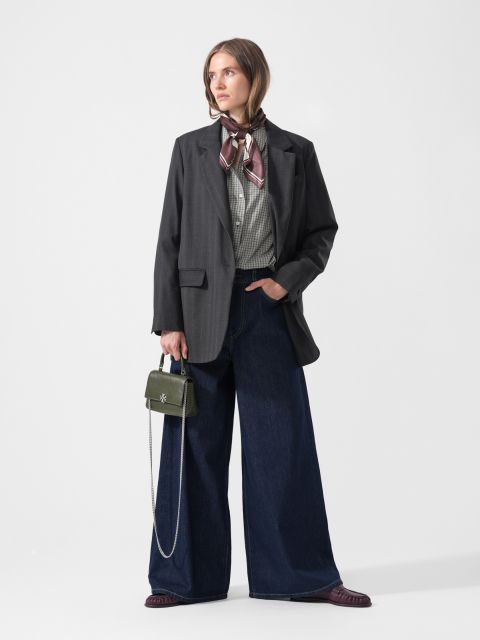 Una donna in jeans a gamba larga, camicia a quadri, blazer scuro e sciarpa, che tiene una piccola borsa verde, è in piedi davanti a un semplice sfondo bianco.