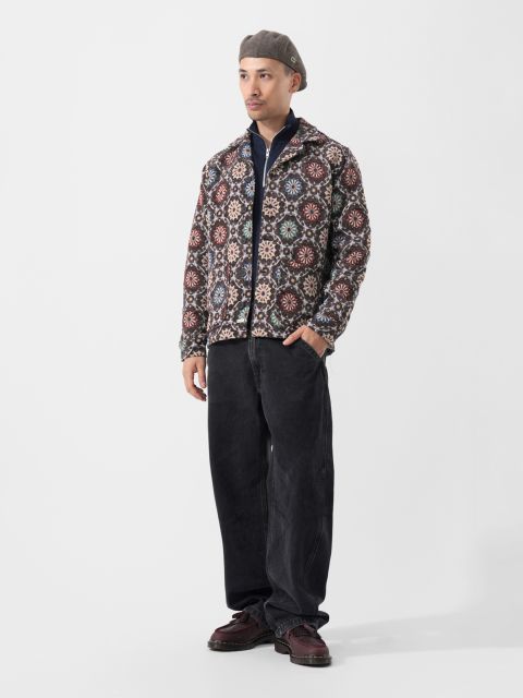 Un uomo con una giacca fantasia, pantaloni neri, scarpe bordeaux e un basco grigio si trova di fronte a un semplice sfondo bianco.