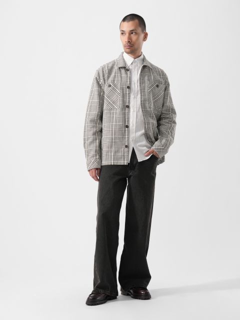 Su un semplice sfondo bianco, un uomo indossa una giacca a quadri sopra una camicia bianca, pantaloni neri larghi e scarpe nere.