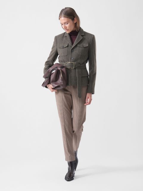 Una donna in blazer con cintura, pantaloni a quadri e stivali tiene in mano una borsa marrone, guarda in basso ed è in piedi davanti a un semplice sfondo bianco.
