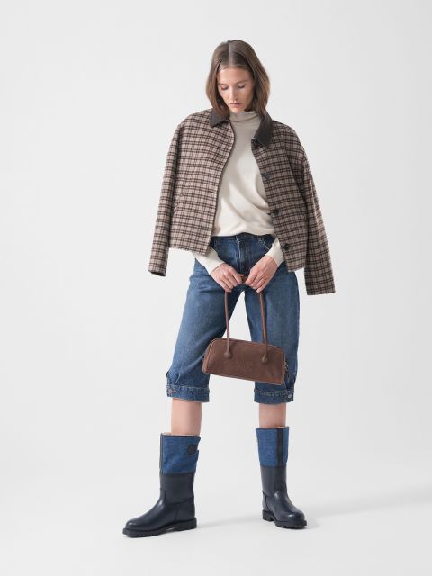 Una donna indossa una giacca a quadri sopra un maglione color crema, pantaloncini di jeans arrotolati e stivali blu navy; tiene in mano una borsa marrone e si trova di fronte a uno sfondo semplice.