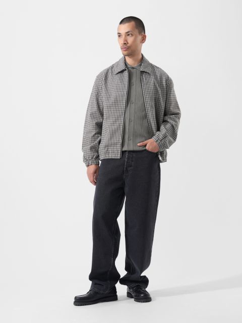 Su uno sfondo bianco semplice, un uomo indossa una giacca a quadri grigia, una camicia grigia, jeans neri larghi e scarpe nere.