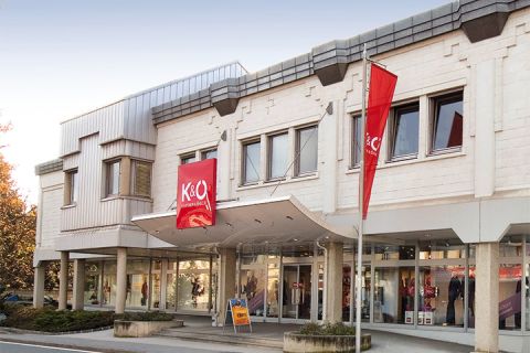 L'edificio moderno con facciata beige ospita i negozi K&Ö Kastner & Öhler e Giga X Sport. Insegne rosse e arancioni sono ben visibili. Grandi finestre e una strada antistante sotto un cielo limpido completano l'immagine.