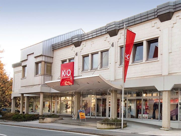 L'edificio moderno con facciata beige ospita i negozi K&Ö Kastner & Öhler e Giga X Sport. Insegne rosse e arancioni sono ben visibili. Grandi finestre e una strada antistante sotto un cielo limpido completano l'immagine.