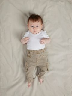 Un bambino sorridente è sdraiato su una coperta bianca e indossa un maglione blu di lana, pantaloni di velluto a coste marroni con tasche e calzini grigi. Il bambino sembra felice e rilassato e ha le braccia leggermente piegate all'altezza dei gomiti.