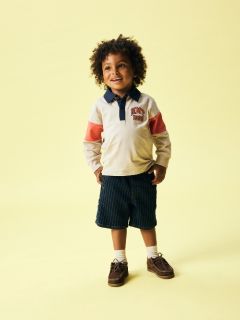 Un ragazzo con i capelli biondi indossa un berretto nero, un pile color crema sopra una camicia a righe, jeans grigi e scarpe da ginnastica. È in piedi di fronte a uno sfondo blu uniforme e guarda in basso con le mani giunte.