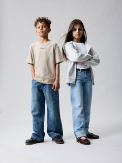 Due bambini piccoli stanno in piedi tenendosi per mano davanti a uno sfondo marrone uniforme. Un bambino indossa una giacca rosa e una gonna, l'altro jeans blu e una camicia blu con la scritta "L'AMORE È BAMBINO". Entrambi guardano in su con curiosità.
