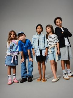 Una bambina con i capelli biondi sorride in piedi davanti a uno sfondo verde, indossando un maglione marrone senza maniche sopra un vestito a fiori, calzini al ginocchio e scarpe marroni.