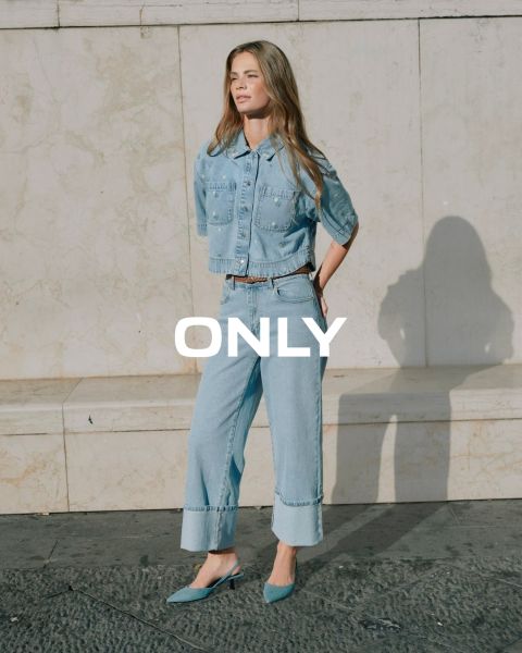 Una giovane donna con un cappotto nero, una camicia a righe blu e una gonna marrone cammina per una strada in città con edifici storici sullo sfondo. La parola "SOLO" è visualizzata in sovrimpressione sull'immagine.