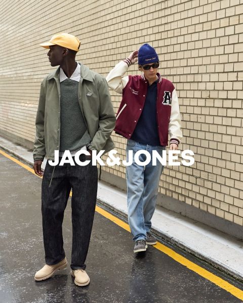 Due uomini in abiti di jeans siedono davanti a uno sfondo semplice. L'uomo davanti si tiene una mano sulla bocca, mentre l'altro è seduto dietro di lui. Al centro c'è il testo "JACK & JONES".