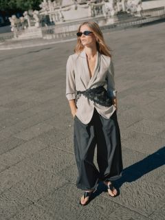 Una donna in un completo marrone oversize con cintura nera e gioielli d'oro cammina con sicurezza attraverso una strada cittadina, portando una grande borsa nera. Sullo sfondo si vedono persone sedute ai tavolini di un caffè.