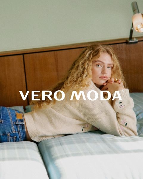 Una donna con gli occhiali e un abito floreale con volant è appoggiata a un muro bianco davanti a uno sfondo piastrellato di blu. La scritta "VERO MODA" appare al centro dell'immagine.