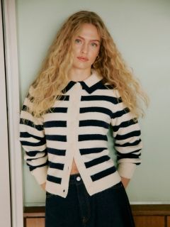 Una donna con i capelli ricci è in piedi accanto a una parete piastrellata. Indossa un cardigan grigio chiaro e soffice, pantaloncini di pelle neri a vita alta e collant neri, con le mani in tasca e un'espressione sicura di sé.