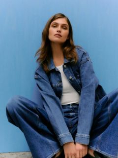 Una persona con lunghi capelli biondi lisci e frangia indossa una giacca di pelle lucida color bordeaux e pantaloni abbinati, appoggiata a un muro di pietra grigio chiaro.