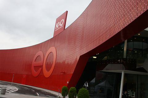 Un edificio moderno con una grande facciata rossa curva con le lettere "eo". Sul tetto si vedono insegne per K&O e Giga Sport. L'ingresso è in vetro, accanto ci sono piante in vaso e in primo piano c'è una curva.