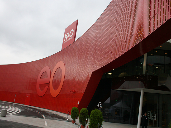 Un edificio moderno con una grande facciata rossa curva con le lettere "eo". Sul tetto si vedono insegne per K&O e Giga Sport. L'ingresso è in vetro, accanto ci sono piante in vaso e in primo piano c'è una curva.