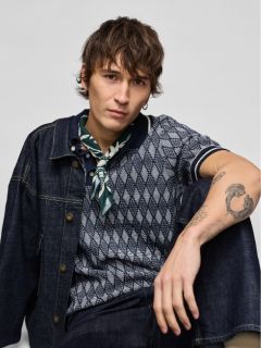 Un giovane uomo in giacca di jeans e jeans è inginocchiato e guarda di lato. Indossa scarpe da ginnastica bianche e ha un tatuaggio visibile sull'avambraccio. Lo sfondo è grigio chiaro uniforme.