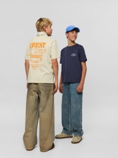 Due ragazzi in jeans e camicie di jeans posano insieme. Uno è seduto su uno sgabello con le mani in grembo e una sciarpa al collo, mentre l'altro ha un braccio sullo sgabello e indossa un berretto.