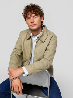 Un giovane uomo che indossa una maglietta nera con la scritta "Original Studio", una collana d'argento, un braccialetto d'argento e un berretto da baseball a righe color crema e nero, è in piedi davanti a uno sfondo semplice grigio chiaro.