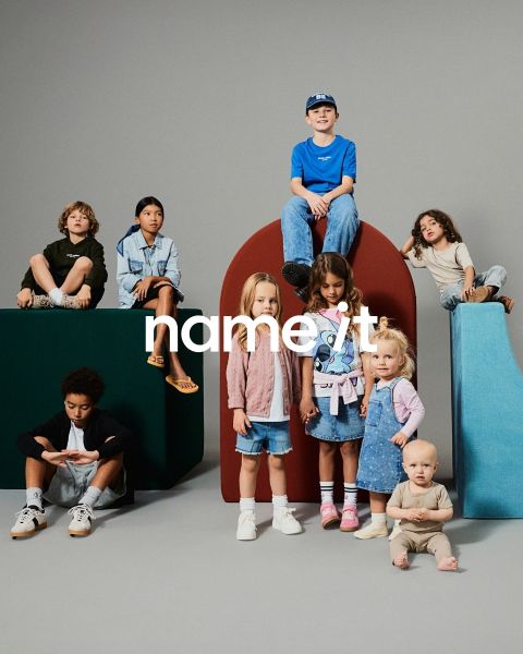 Un gruppo di otto bambini diversi è in piedi insieme su uno sfondo grigio, tutti indossano abiti casual coordinati in colori neutri e pastello. Il testo "name it" appare al centro dell'immagine.