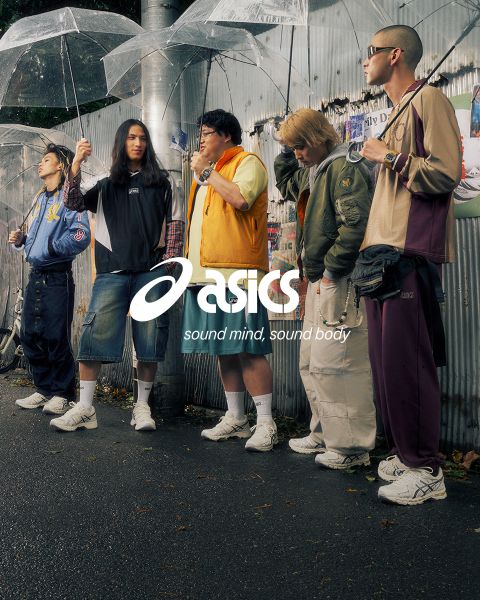 Due persone in abiti alla moda sono in piedi con delle biciclette su una strada acciottolata. Uno indossa una camicia blu e pantaloni bianchi, l'altro una giacca e pantaloni scuri. La scena è soleggiata e sullo sfondo si vedono vecchi edifici. Il logo Asics è visibile.