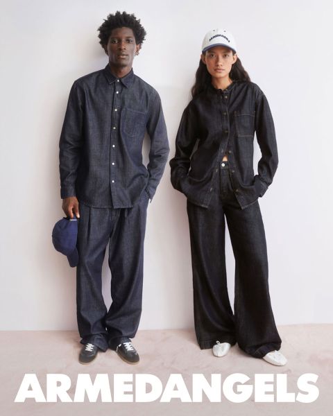 Due giovani adulti in abbigliamento di jeans stanno insieme all'aperto, uno con il braccio alzato, l'altro con una valigia coperta di adesivi. In basso, in grassetto e scritta bianca, c'è la scritta "ARMEDANGELS".