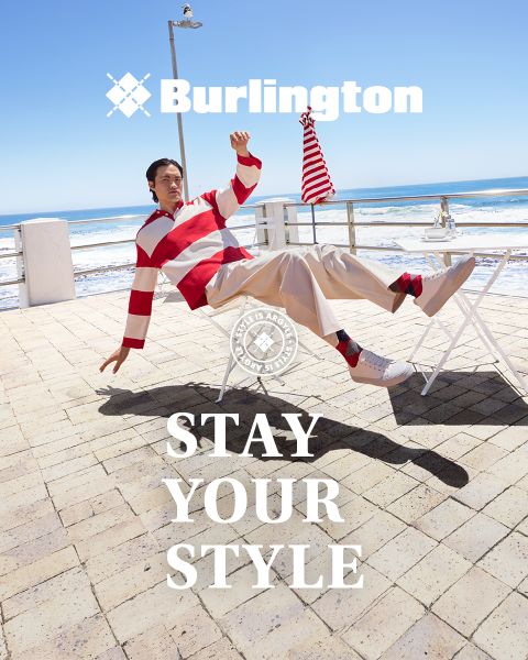 Un uomo e una donna in abiti eleganti posano con sicurezza su una panchina bianca davanti a un muro verde con i testi "Burlington" e "Stay Your Style" sopra e sotto di loro.
