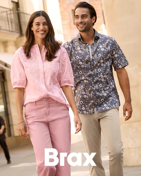 Una donna con un cappotto fantasia e un maglione rosso è in piedi accanto a un uomo con un cappotto invernale blu navy e pantaloni beige. Sono appoggiati a un muro di pietra all'aperto. La parola "Brax" appare nella parte inferiore dell'immagine.