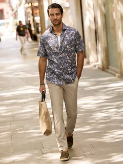 Un uomo con capelli scuri corti e la barba è appoggiato a una ringhiera di pietra all'aperto. Indossa una giacca a quadri rossi e bianchi sopra un maglione color crema e jeans blu. Sullo sfondo si vedono edifici urbani.