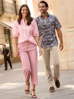 Una donna con un cappotto di pelliccia fantasia e un top rosso sorride accanto a un uomo con una giacca invernale blu navy e un maglione beige. Sono in piedi all'aperto vicino a un muro di pietra e la donna tiene in mano una borsa marrone.