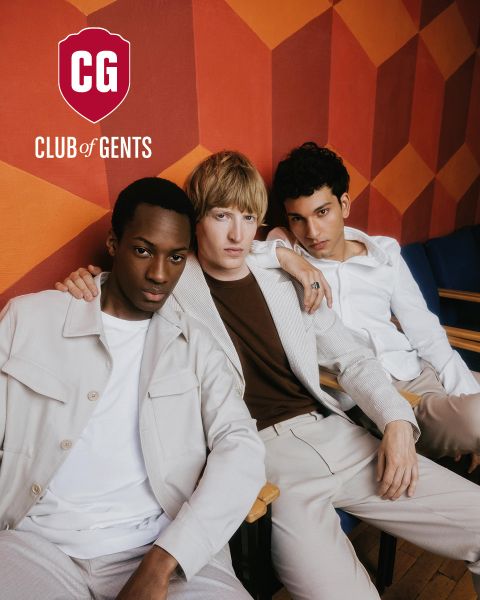 Due uomini posano per una foto di moda; uno indossa un abito gessato marrone chiaro e una camicia scura, mentre l'altro è seduto accanto a lui con una camicia nera. Lo sfondo è leggermente strutturato, con il logo del "CG Club of Gents" sopra di loro.