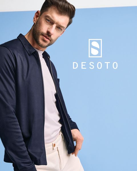 Un uomo con una camicia a maniche lunghe fantasia e pantaloni scuri è in piedi davanti a uno sfondo grigio. Guarda di lato con le mani in tasca. La scritta "DESOTO" e il logo DESOTO sono visibili nella parte inferiore.