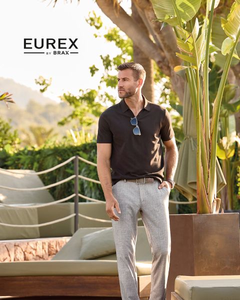 Un uomo è in piedi all'aperto vicino all'erba alta, indossa un maglione beige, un gilet imbottito blu navy e pantaloni marroni e guarda di lato. Il logo "EUREX by BRAX" appare sul lato destro. Il cielo è nuvoloso.