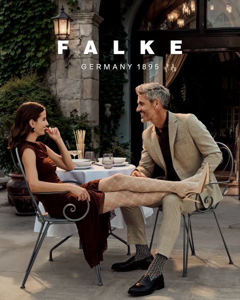 Una modella in un blazer color castagna, guanti marroni, collant verdi, una gonna grigia con spacchi alti e stivaletti marroni posa davanti a uno sfondo geometrico. Il testo recita "FALKE GERMANY 1895".