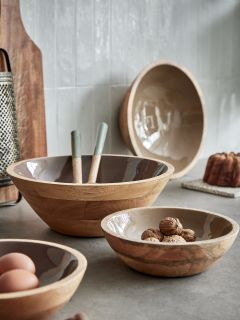 Su un tavolo da cucina ci sono tre ciotole di legno di diverse dimensioni. In una ciotola ci sono noci, in un'altra due uova e nella più grande ci sono utensili. Sullo sfondo si vedono una grattugia, un tagliere e una ciotola fissata al muro.