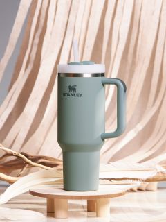 Un bicchiere termico Stanley verde con manico e coperchio con cannuccia è posizionato su un piccolo piedistallo di legno. Lo sfondo è composto da un tessuto beige drappeggiato e rami decorativi.