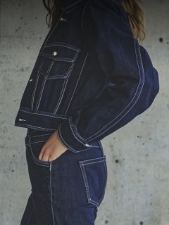 Una donna con un cappello bianco, una maglietta bianca e jeans blu posa con il braccio teso in una stanza moderna e bianca con dei fiori sopra.