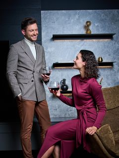 Un uomo in giacca e una donna in un lungo abito bordeaux sorridono tenendo in mano bicchieri di vino rosso in un soggiorno moderno ed elegante con scaffali decorativi e una clessidra al muro.