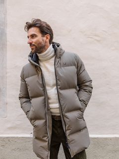 Un uomo con capelli castani corti e barba indossa un lungo piumino marrone chiaro sopra un dolcevita bianco. È in piedi accanto a un muro chiaro e guarda a sinistra, con le mani nelle tasche del cappotto.